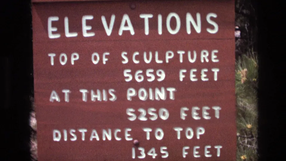 1974 8mm frame at Mount Rushmore showing an elevation sign reading top of sculpture 5659 feet, at this point 5250 feet, and distance to top 1345 feet.