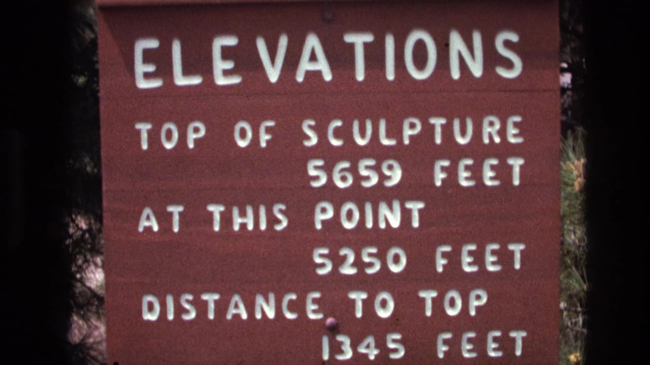 Close-up 1974 South Dakota 8mm frame of a red wooden sign reading elevations, with white text listing the sculpture top, current point, and distance to top.