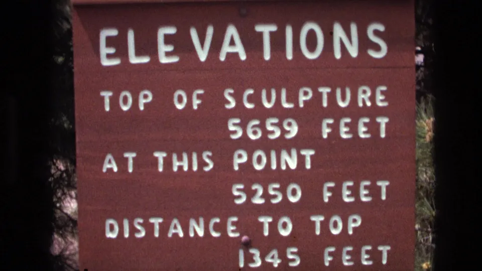 Close-up 1974 South Dakota 8mm frame of a red wooden sign reading elevations, with white text listing the sculpture top, current point, and distance to top.