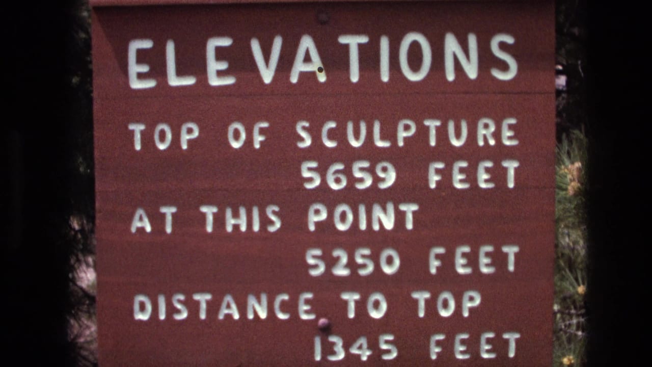 Close visitor-shot frame of the South Dakota elevations sign, showing the top of sculpture measurement, current elevation, and distance to top in white letters.