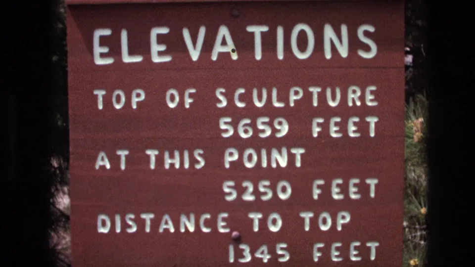 Close visitor-shot frame of the South Dakota elevations sign, showing the top of sculpture measurement, current elevation, and distance to top in white letters.