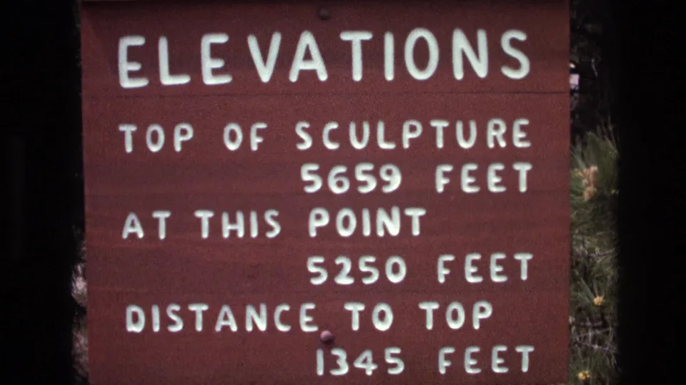 Handheld 8mm close-up of the red South Dakota elevations sign, with the white measurement text still legible and vegetation visible at the margins.