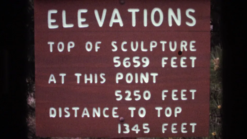 Close 8mm visitor-shot frame of the Mount Rushmore elevations sign, with white text on brown wood and evergreen foliage visible at the edge.