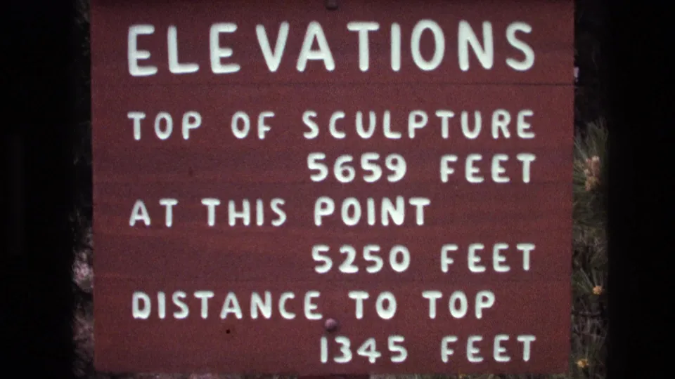 Final close 8mm frame of the Mount Rushmore elevations sign, with legible white measurement lines and wooded surroundings at the margins.