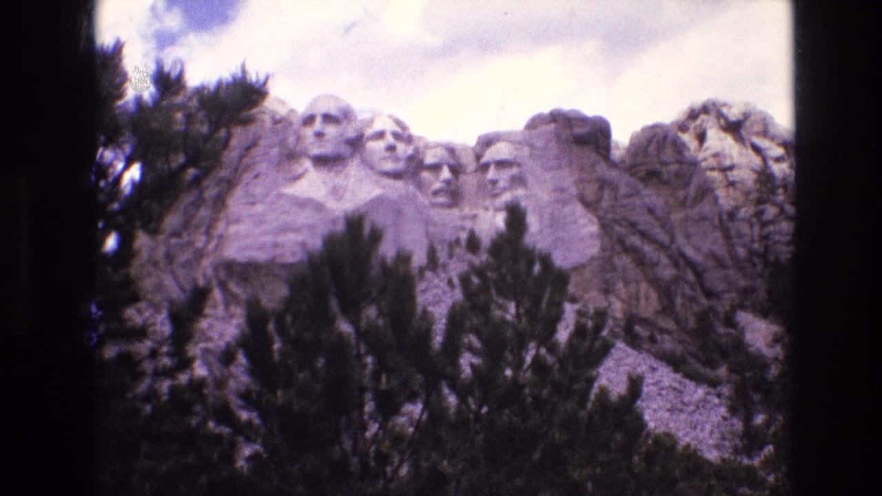1974 8mm frame of Mount Rushmore National Memorial in South Dakota, with the carved presidential faces partly framed by pine trees and surrounding granite.