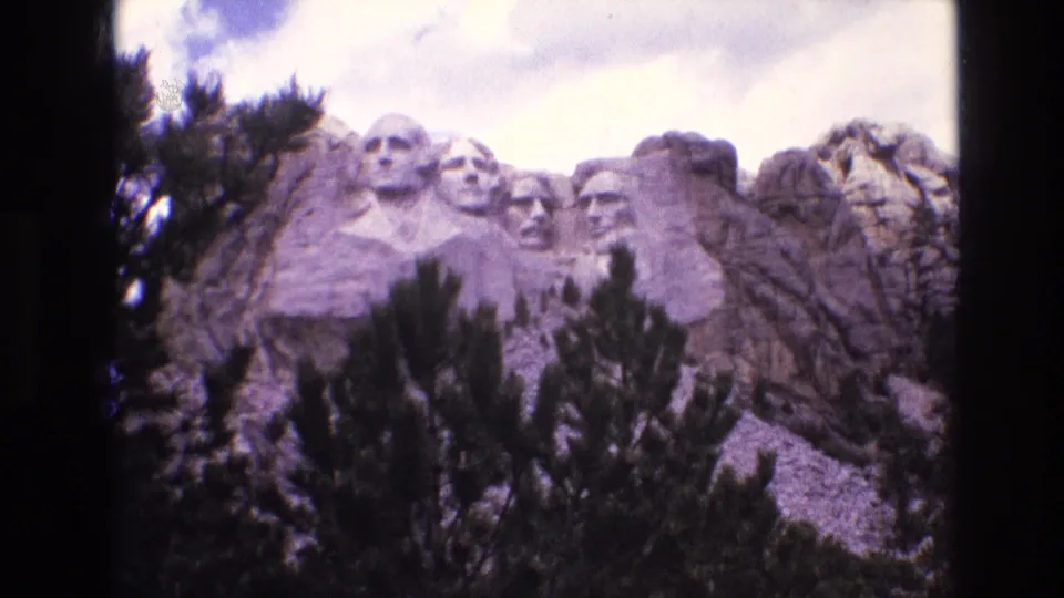 1974 8mm frame of Mount Rushmore National Memorial in South Dakota, with the carved presidential faces partly framed by pine trees and surrounding granite.
