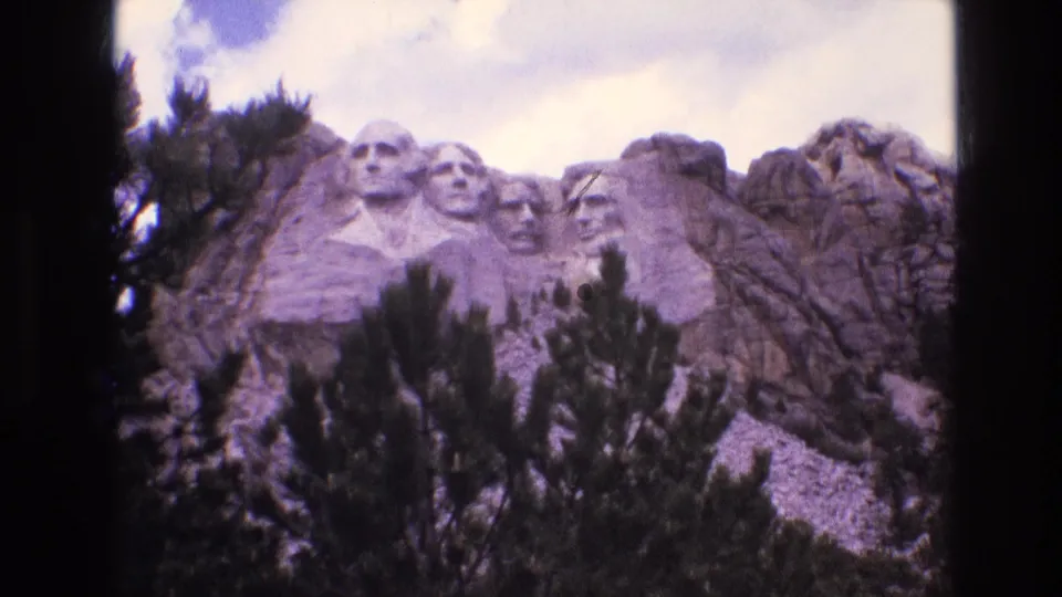 1974 8mm frame of Mount Rushmore National Memorial, with the granite faces partially obscured by dark pine branches and soft, faded color in the sky and rock.