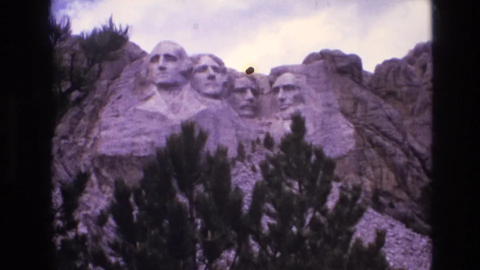 1974 8mm home-movie frame of Mount Rushmore in South Dakota, showing the carved presidential faces close in frame with pine branches, granite formations, soft focus, and a small dust mark.