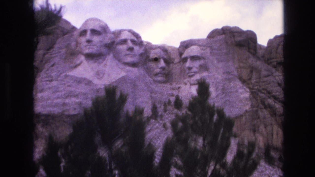 1974 8mm travel frame of Mount Rushmore in South Dakota, showing the four presidential carvings behind evergreen branches and soft purple film tint.