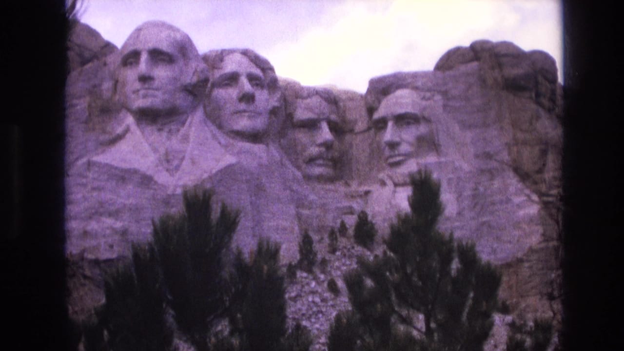 1974 8mm frame from South Dakota showing Mount Rushmore's carved presidential heads, foreground evergreens, cloudy sky, and visible home-movie frame edges.