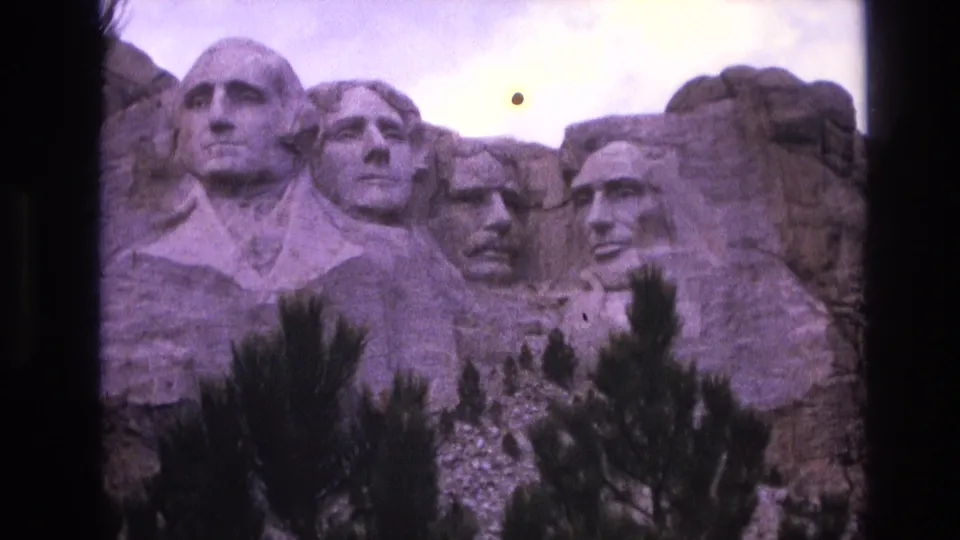 1974 8mm South Dakota travel frame showing Mount Rushmore from below, with Washington prominent, other carved heads receding, and visible dust on the film.