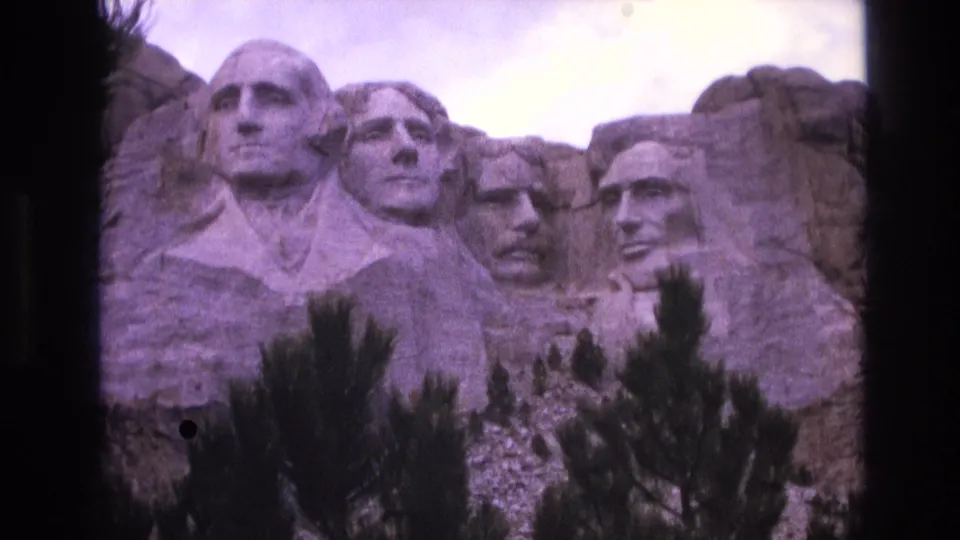 1974 home-movie frame of Mount Rushmore in South Dakota, holding on the presidential carvings as foreground trees obscure part of the lower monument.