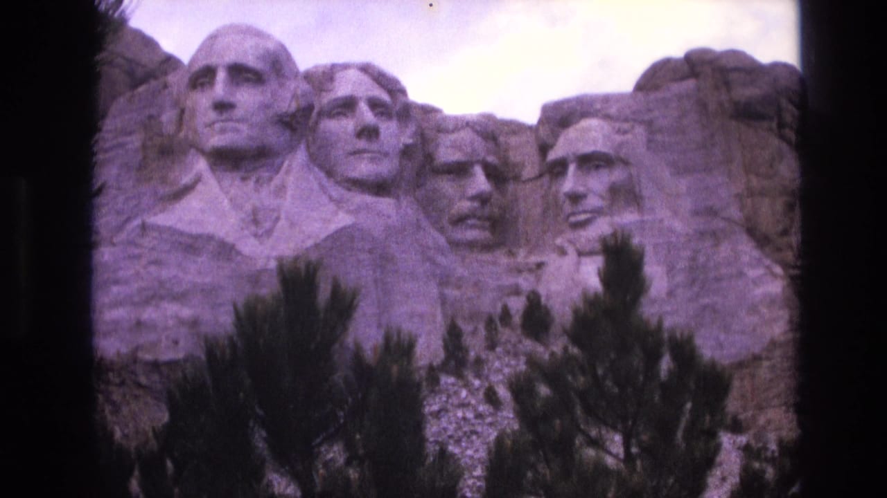 1974 8mm view of Mount Rushmore National Memorial in South Dakota, with the four carved presidential faces partly framed by foreground pine trees and dark film borders.