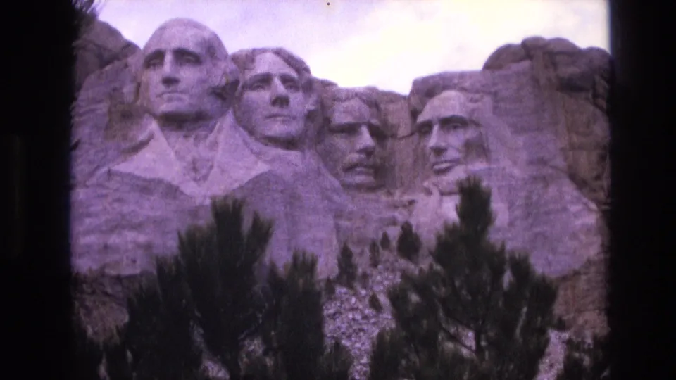 1974 8mm view of Mount Rushmore National Memorial in South Dakota, with the four carved presidential faces partly framed by foreground pine trees and dark film borders.