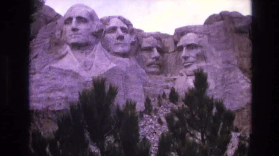 Handheld 1974 8mm frame showing Mount Rushmore's carved presidential faces with pine trees in the foreground, pale sky above, and visible dark film-frame borders.