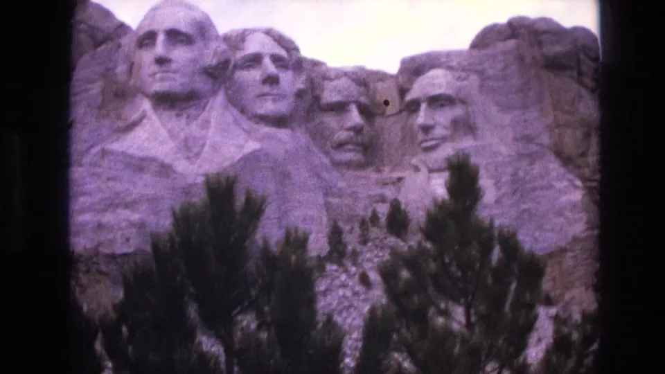 1974 South Dakota home-movie frame of Mount Rushmore, with soft focus, purple color shift, and pine trees rising across the lower portion of the monument.
