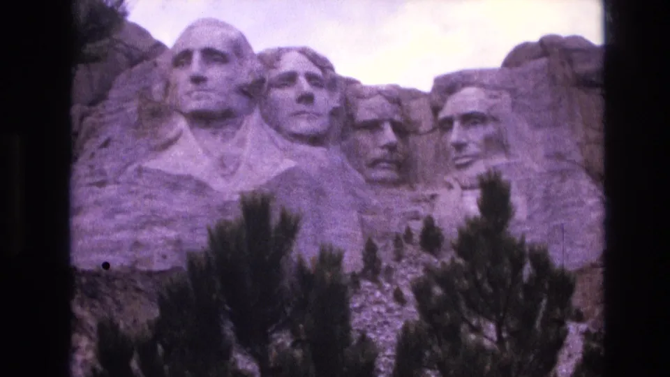 Final 1974 8mm frame of Mount Rushmore National Memorial, showing the carved faces behind dark pine silhouettes with grain, purple tint, and rounded film-frame edges.