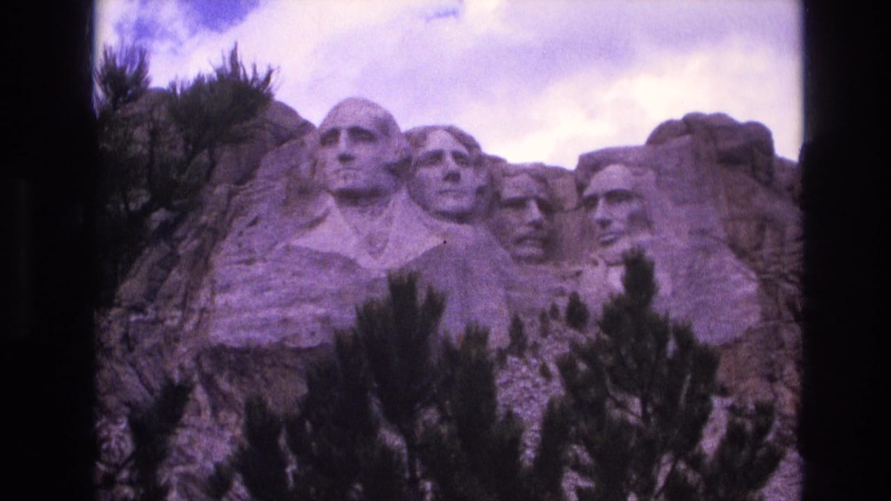 1974 visitor-shot 8mm view of the Mount Rushmore granite sculpture, with the four presidential heads visible above dark pine branches under partly cloudy sky.