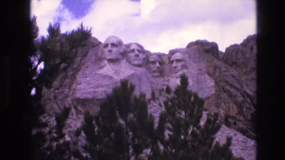 1974 8mm frame of Mount Rushmore in the Black Hills, showing the presidential carvings across the granite face with dark pine foliage covering the lower foreground.