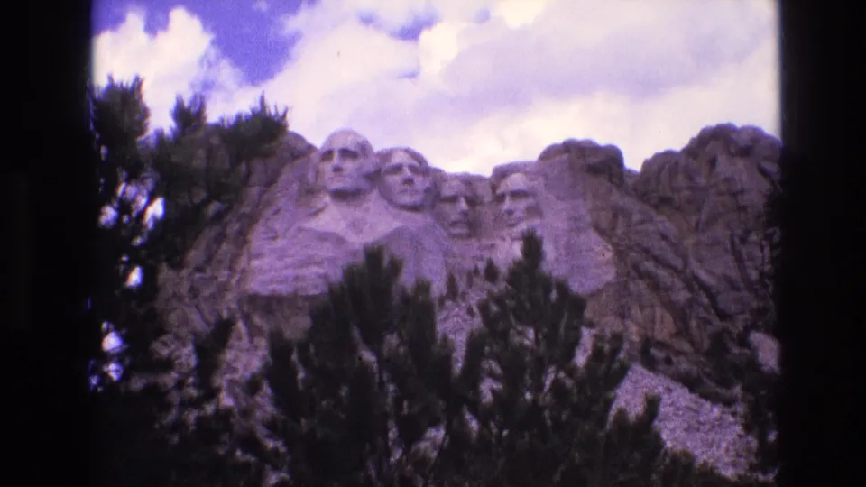Final 1974 home-movie frame of Mount Rushmore National Memorial, with the granite presidential heads centered behind pine branches and a bright partly cloudy sky.