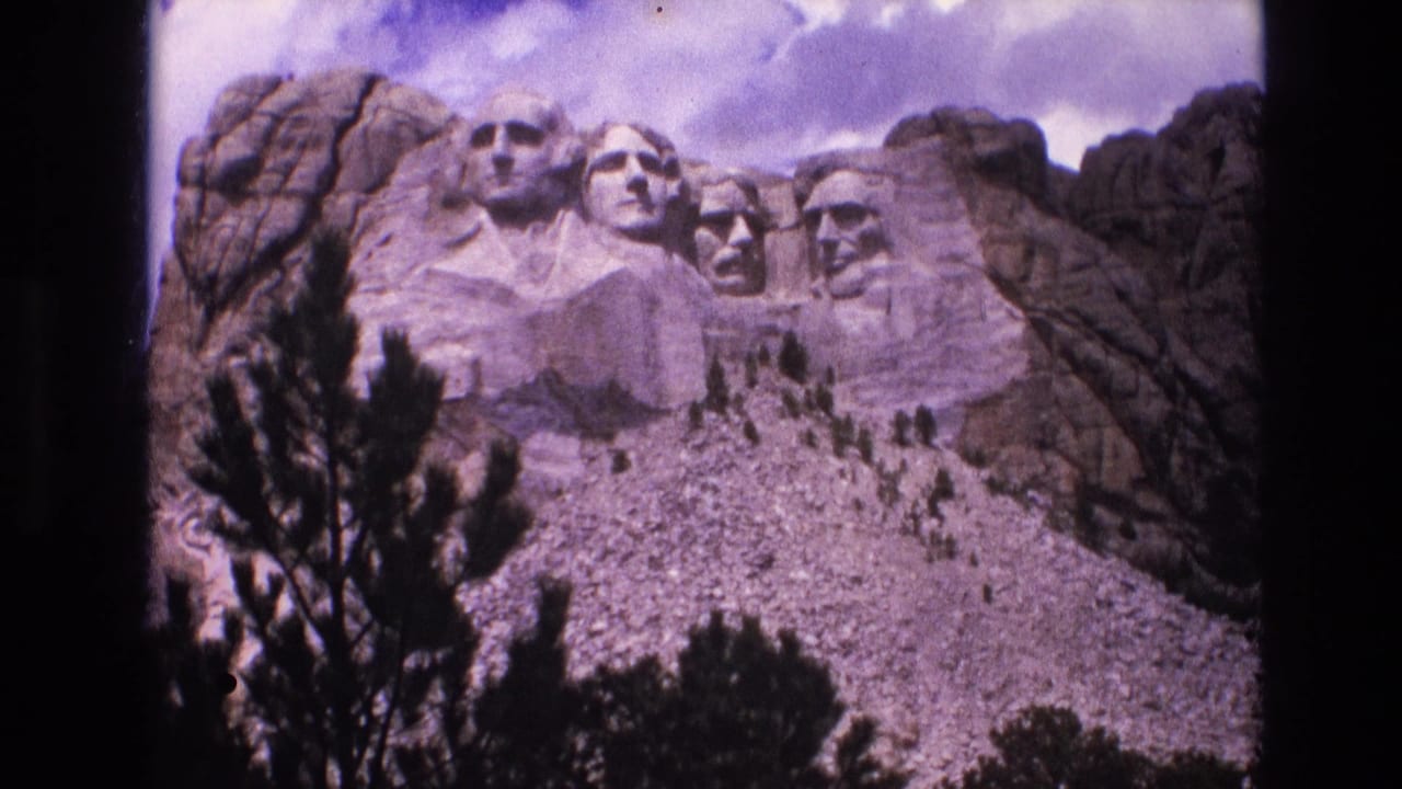 Mount Rushmore presidential sculptures seen above evergreen branches and rocky slope.