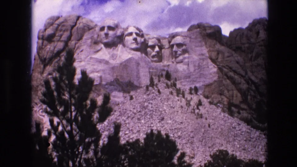 1974 8mm frame of Mount Rushmore in South Dakota, with the presidential heads above a pale rocky slope, dark evergreen branches, and cloudy sky.