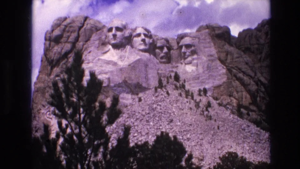 1974 8mm view of Mount Rushmore from a low visitor angle, showing the granite presidential faces, foreground evergreens, rocky hillside, and clouded sky.