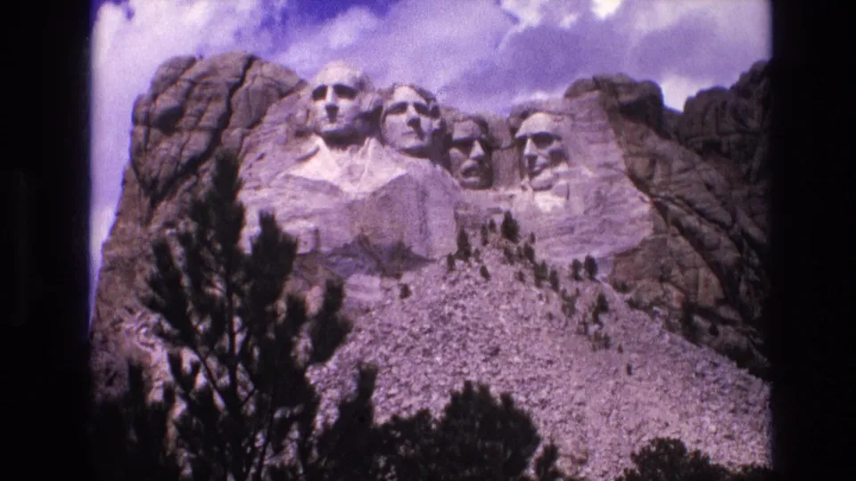 1974 8mm frame showing Mount Rushmore’s carved presidential heads against granite rock, with dark evergreen branches in the lower foreground and a purple-tinted sky.