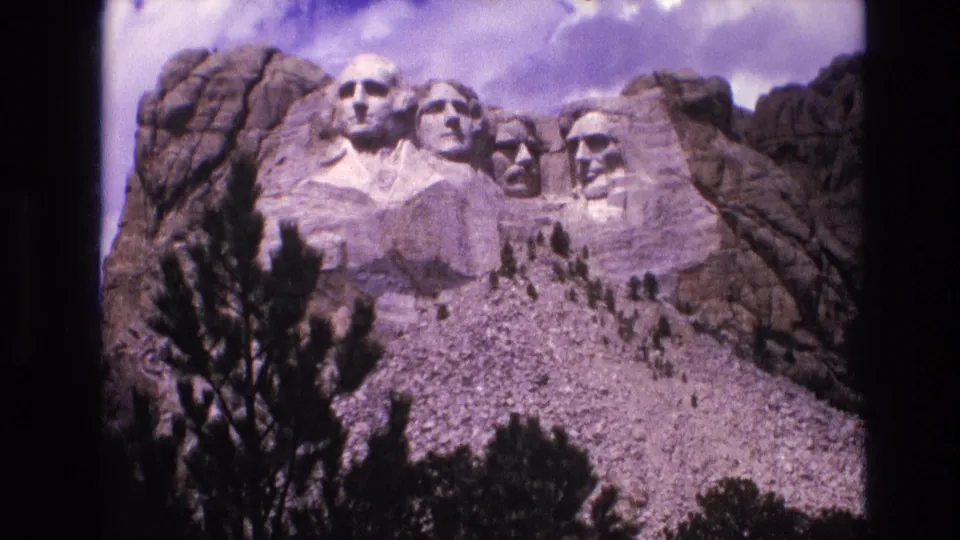 1974 8mm frame of the Mount Rushmore sculptures in South Dakota, with the granite hillside, scattered evergreens, cloudy sky, and visible home-movie frame edge.
