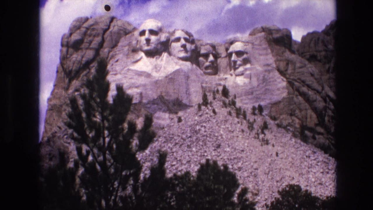 1974 8mm frame of Mount Rushmore in South Dakota, viewed from below with pine trees in the foreground and a partly cloudy sky above.