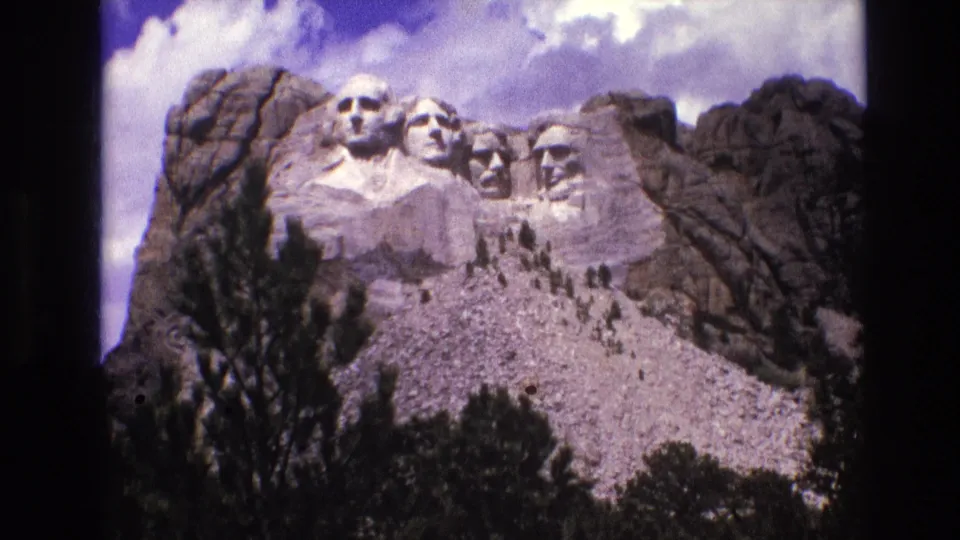 1974 8mm frame showing Mount Rushmore from a low visitor vantage, with dark pine trees at the bottom and rock formations around the carved faces.