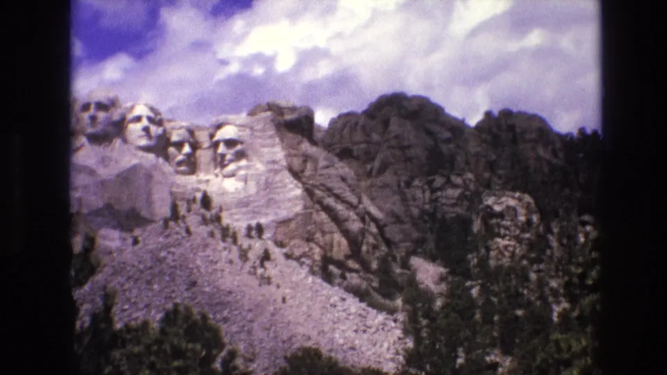 1974 8mm frame with Mount Rushmore shifted toward the left, showing more surrounding rock, trees, and bright clouds over the memorial.