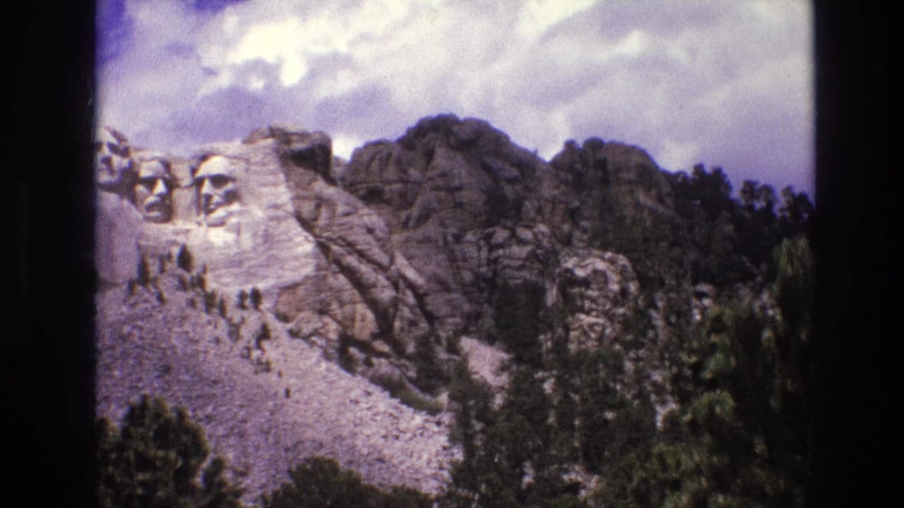 1974 8mm home-movie frame showing Mount Rushmore at the left edge, with granite mountain ridges, pine forest, cloudy sky, and dark film borders.