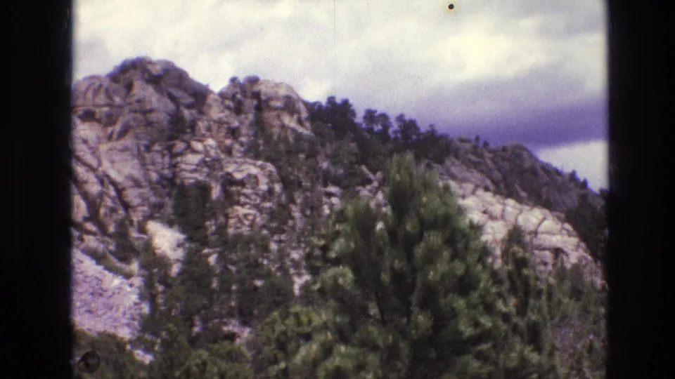1974 8mm frame from the Mount Rushmore area showing rocky granite outcrops, scattered pines, and a cloudy sky as the camera pans away from the carved faces.