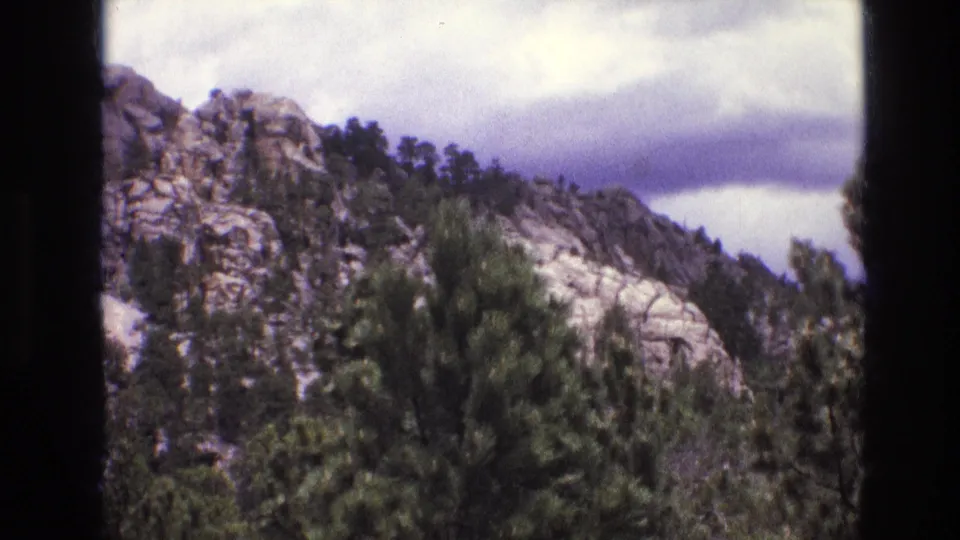 1974 South Dakota home-movie frame showing pine trees in the foreground, pale granite ridges in the background, cloudy sky, and dark film borders from the scan.