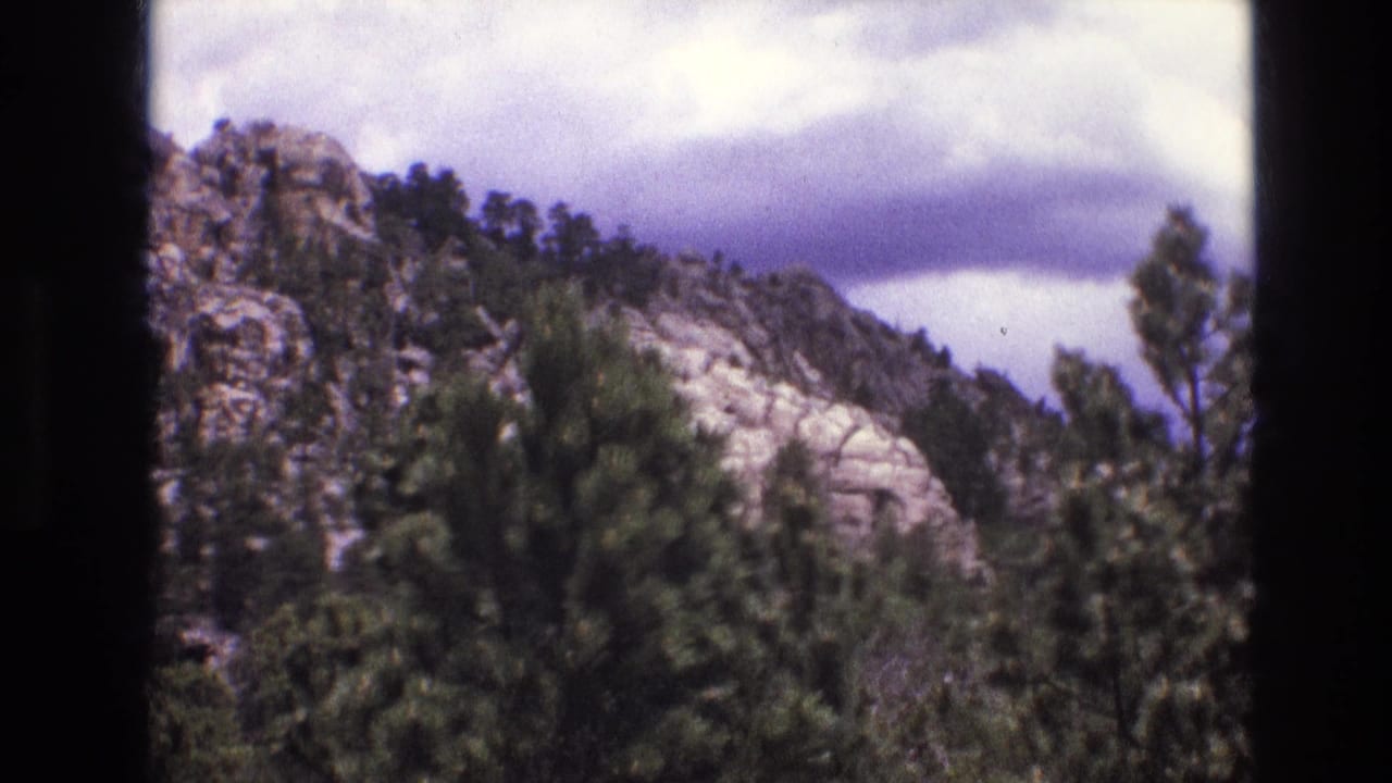 1974 South Dakota 8mm frame showing a rocky mountain slope covered with evergreen trees beneath a cloudy sky.