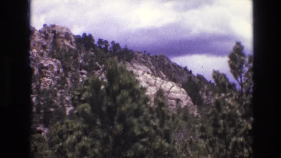 1974 South Dakota 8mm frame showing a rocky mountain slope covered with evergreen trees beneath a cloudy sky.