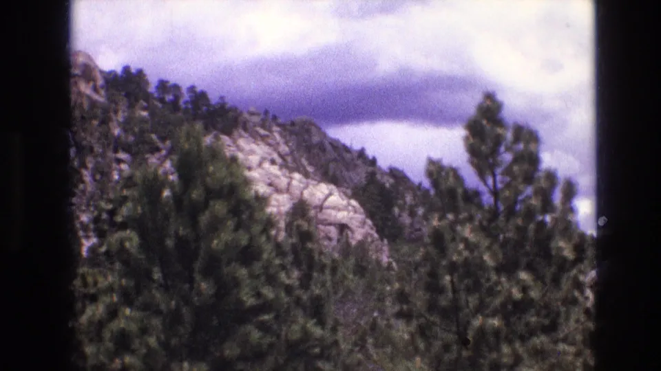 1974 South Dakota home-movie frame with evergreen trees in front of a pale rocky hillside and layered clouds.
