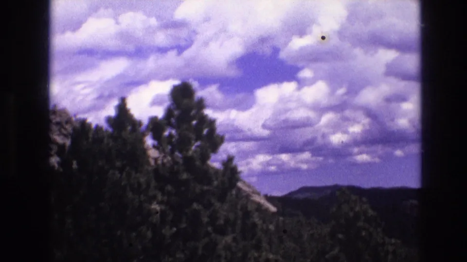 1974 South Dakota 8mm frame looking past dark evergreens toward distant ridges under a broad cloud-filled sky.