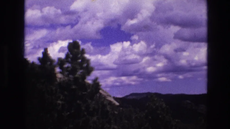 1974 South Dakota home-movie frame showing evergreen trees in the foreground with distant hills beneath a mostly cloudy sky.