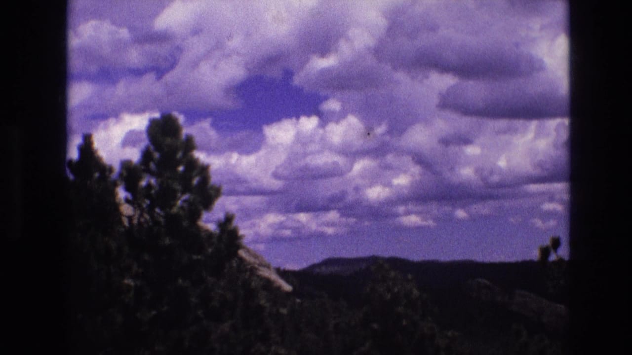 1974 South Dakota 8mm frame showing a cloud-filled blue-purple sky above silhouetted pine trees, low ridges, and dark foreground vegetation.