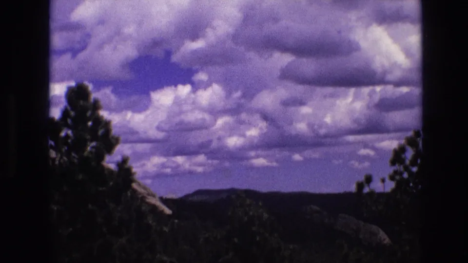 1974 South Dakota home-movie frame with a tall dark pine shape at left, layered clouds overhead, and a low ridge line across the horizon.