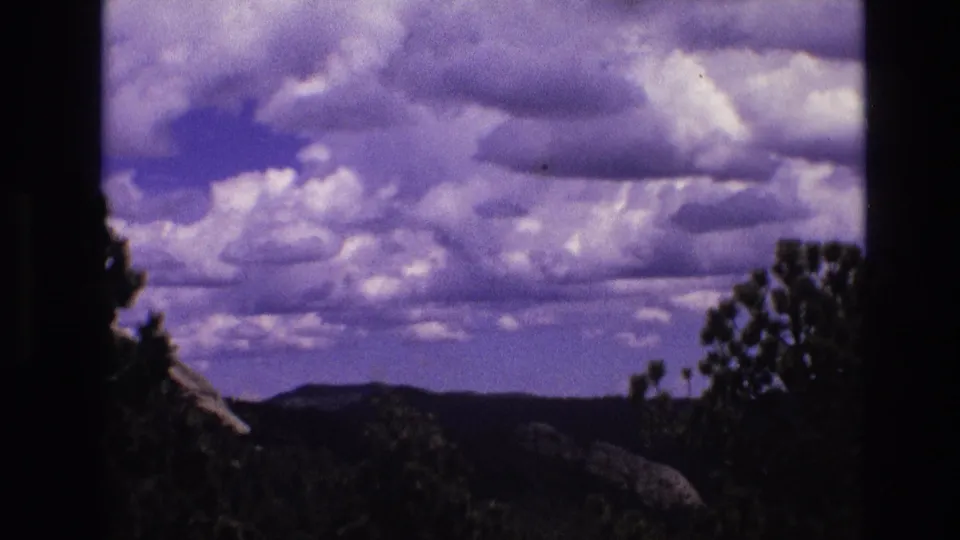 1974 South Dakota 8mm frame emphasizing a broad clouded sky, low wooded horizon, and dark tree and shrub silhouettes at the edges.