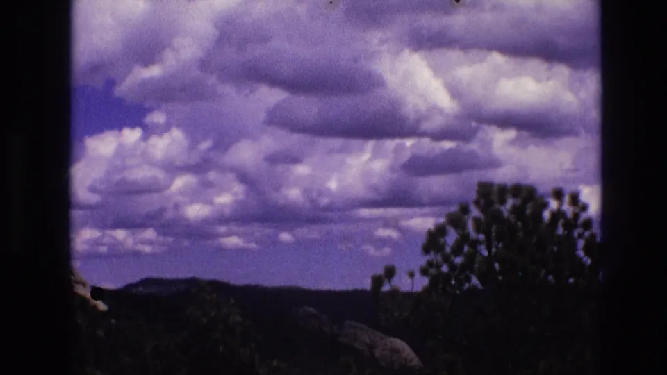1974 South Dakota 8mm frame showing layered clouds over distant wooded hills, with dark pine and shrub silhouettes framing the lower right and left edges.