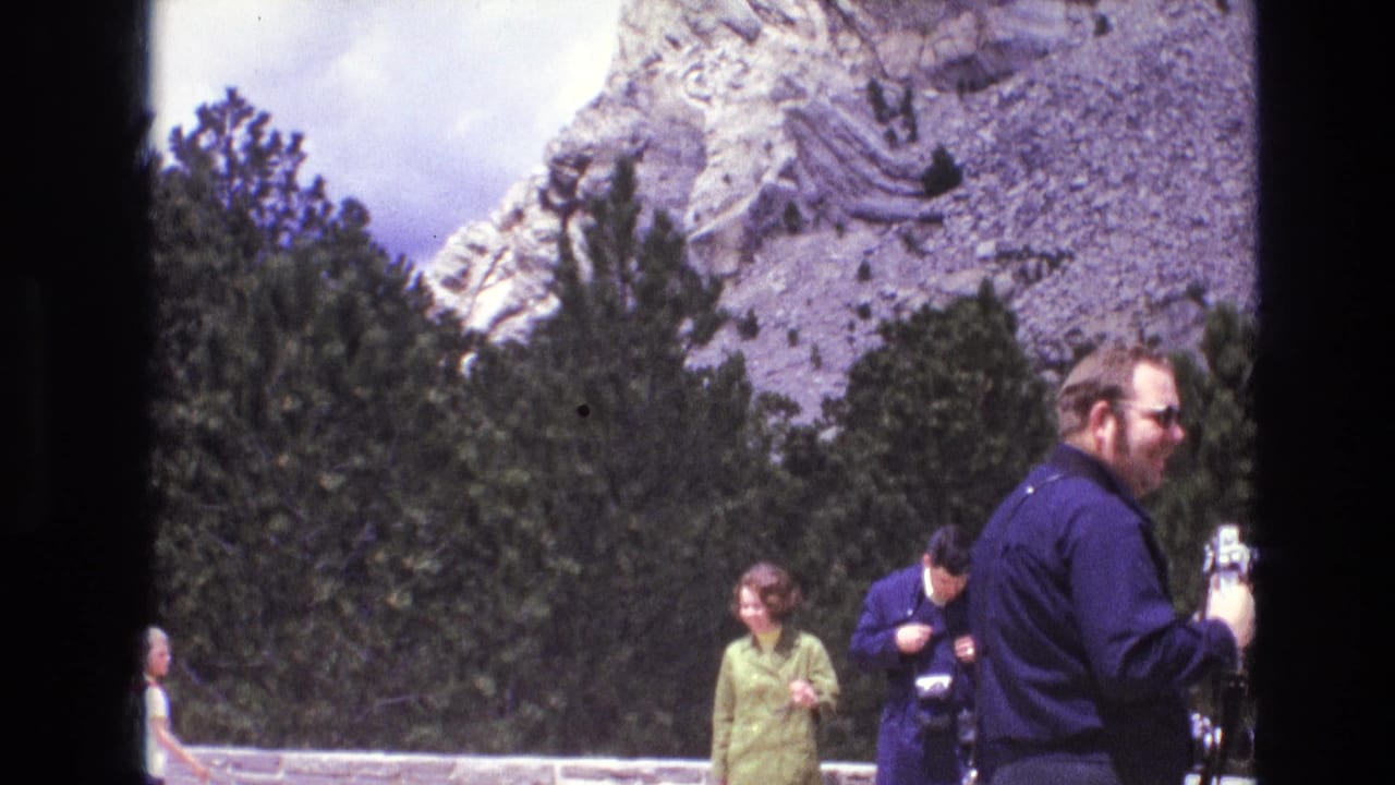 1974 South Dakota 8mm frame showing tourists at a scenic overlook with cameras, evergreen trees, and a carved mountain face behind them.