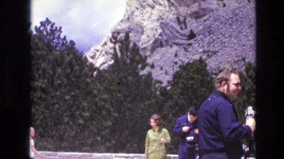 1974 South Dakota 8mm frame showing tourists at a scenic overlook with cameras, evergreen trees, and a carved mountain face behind them.