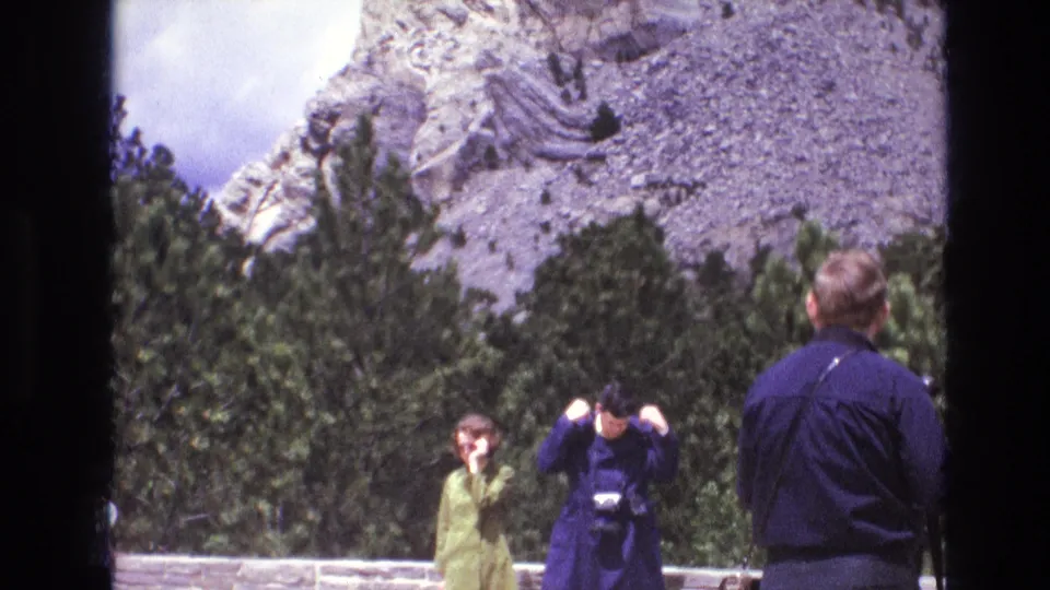 1974 South Dakota home-movie frame with visitors near a low stone wall, one raising a camera, and a carved mountain surface partly visible above the trees.