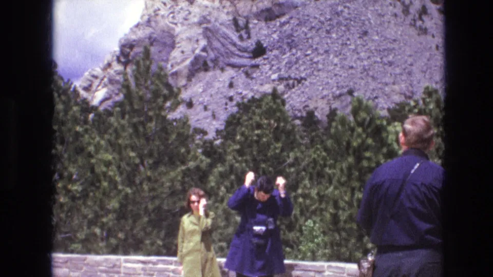 1974 South Dakota 8mm frame showing tourists at a public overlook, with a woman in green, a camera user in dark clothing, trees, and carved rock in the background.