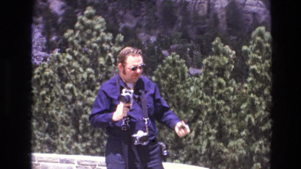 1974 South Dakota home-movie frame focused on a man in sunglasses and a blue shirt carrying cameras, with evergreen trees and rock formations behind him.