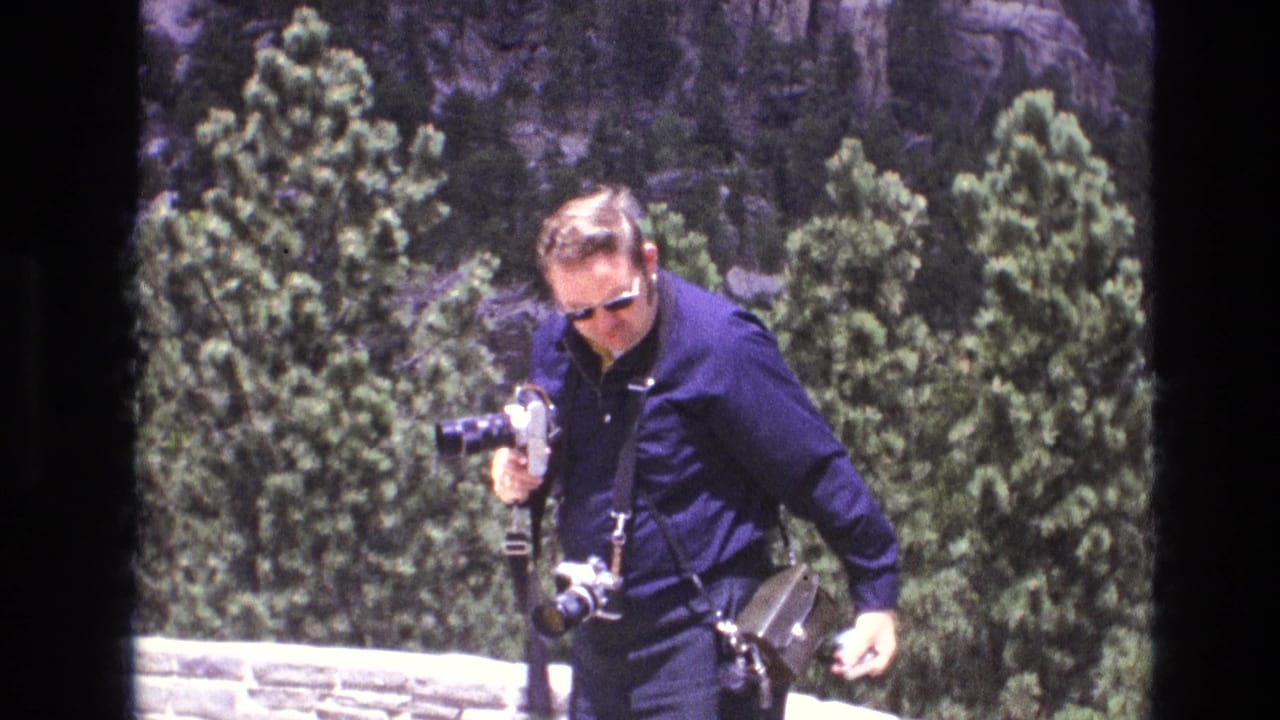 In South Dakota 1974 home-movie footage, a man in a dark jacket and sunglasses holds multiple cameras while standing before evergreen trees and a rocky cliff face.